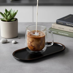 Peut inclure: Une tasse en verre à double paroi remplie de café, avec du lait versé. La tasse est posée sur un plateau ovale noir. Une petite plante succulente dans un pot beige et une pile de livres sont en arrière-plan.