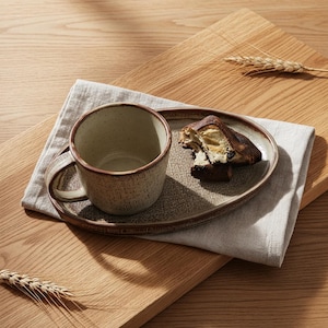 Peut inclure: Une tasse en céramique et un plateau ovale assorti, avec une finition texturée et mouchetée et une bordure marron. Un morceau de pain est posé sur le plateau. L'ensemble est posé sur une serviette en lin et une planche en bois.