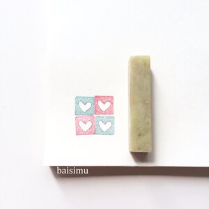 Può includere: Un timbro rettangolare verde con quattro quadrati, ognuno contenente un cuore bianco. I cuori sono in un motivo rosa e blu. Il timbro è accanto a un pezzo di carta bianca con la parola "baisimu" scritta su di esso.