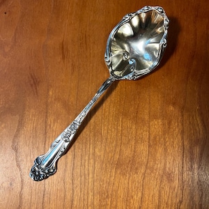 May include: A silver serving spoon with an ornate floral design on the handle and a scalloped bowl. The spoon is made of a shiny, reflective metal and is set against a warm, wooden background. This spoon is ideal for serving food.