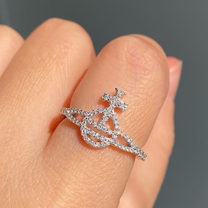 May include: A silver ring featuring a planet and cross design, encrusted with small, sparkling stones. The ring is worn on a finger, showcasing its intricate details and overall elegance. The ring's design is a unique and eye-catching piece of jewelry.