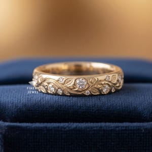 May include: A gold ring with a floral and leaf design, set with multiple small diamonds. The ring is displayed on a dark blue velvet surface, with a blurred gold background.