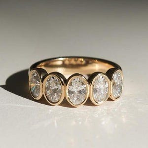 Puede incluir: Un anillo de oro con seis diamantes de talla ovalada dispuestos en fila. El anillo tiene una banda de oro amarillo pulido. Los diamantes son claros y brillantes. El anillo se muestra sobre una superficie blanca con iluminación suave.