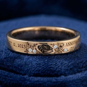 May include: A gold band ring featuring an engraved Bible icon, floral motifs, and three small inset diamonds. The exterior is inscribed with the text OCT 12, 2023 and ANNIVERSARY, along with a small symbol of two interlocking wedding rings.
