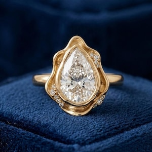 May include: A gold ring featuring a large pear-shaped center stone surrounded by a fluid, organic-shaped metal setting. Small, round accent stones are embedded along the edges of the gold frame, creating a textured, layered appearance.