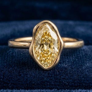 May include: A gold ring featuring a marquise-cut yellow gemstone set in a smooth, organic-shaped bezel setting. The band is polished gold, and the central stone displays a faceted surface with a bright, warm yellow hue and high clarity.