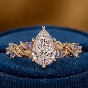 May include: A gold ring with a pear-shaped diamond center stone. The band features seashell and leaf designs, with small blue gemstones. The ring is set against a blue velvet background.