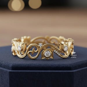 以下が含まれることがあります： 金色のリングで、装飾的な花の模様が施されています。リングには、透明で丸い宝石と繊細なスクロールワークがいくつかあります。リングは濃い青色の表面に置かれ、背景はぼやけています。