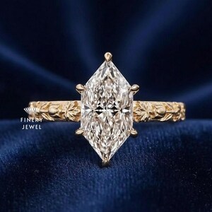 May include: A marquise-cut diamond engagement ring with a gold band. The diamond is set with six prongs and the band features a floral design. The ring is displayed on a dark blue velvet surface. The text "FINERY JEWEL" is visible.