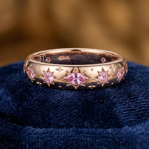 May include: Rose gold ring featuring a celestial design. The ring is adorned with pink gemstones set within star and diamond-shaped cutouts. The band has a smooth, polished finish, and the ring is displayed on a dark blue velvet surface.