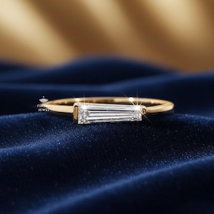 May include: A gold band ring with a rectangular, clear gemstone. The ring is set on a dark blue velvet surface. The gemstone is a baguette cut, and the ring has a simple, elegant design.
