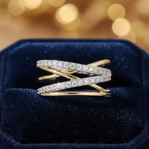 K&ouml;nnte beinhalten: Ein goldfarbener Ring mit einem Kreuzmuster, besetzt mit kleinen, funkelnden wei&szlig;en Edelsteinen. Der Ring befindet sich auf einem dunkelblauen Samthintergrund mit einem verschwommenen, goldenen Bokeh-Effekt.