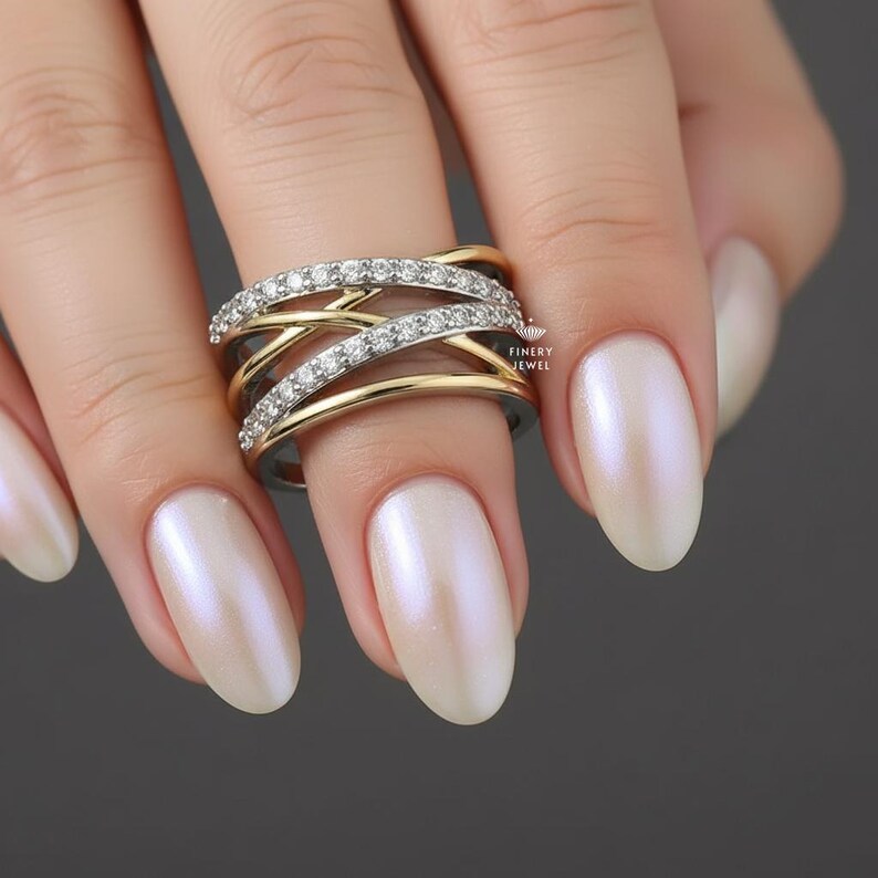 K&ouml;nnte beinhalten: Ein gold-silberner Ring mit einem Kreuzmuster, verziert mit kleinen, funkelnden Edelsteinen. Der Ring befindet sich an einem Finger mit polierten, perlmuttfarbenen N&auml;geln. Das Finery Jewel-Logo ist sichtbar.