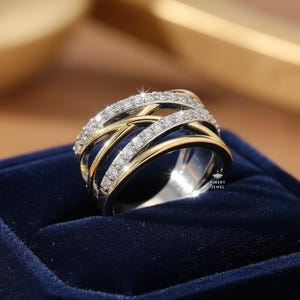 K&ouml;nnte beinhalten: Ein zweifarbiger Ring mit einem silbernen Band und goldenen Akzenten. Der Ring verf&uuml;gt &uuml;ber mehrere miteinander verwobene B&auml;nder, die jeweils mit kleinen, funkelnden Diamanten besetzt sind. Der Ring ist auf einer dunkelblauen Samtoberfl&auml;che platziert, was ein luxuri&ouml;ses Gef&uuml;hl erzeugt.