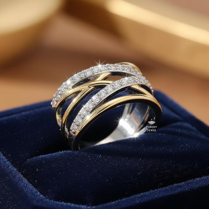 K&ouml;nnte beinhalten: Ein zweifarbiger Ring mit einem silbernen Band und goldenen Akzenten. Der Ring verf&uuml;gt &uuml;ber mehrere miteinander verwobene B&auml;nder, die jeweils mit kleinen, funkelnden Diamanten besetzt sind. Der Ring ist auf einer dunkelblauen Samtoberfl&auml;che platziert, was ein luxuri&ouml;ses Gef&uuml;hl erzeugt.