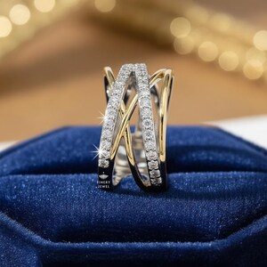 K&ouml;nnte beinhalten: Ein zweifarbiger Ring mit einem Kreuzmuster. Der Ring hat ein Band aus kleinen, klaren Steinen, die in einem silberfarbenen Metall gefasst sind, mit goldfarbenen Akzenten. Der Ring wird auf einer dunkelblauen Samtoberfl&auml;che pr&auml;sentiert.