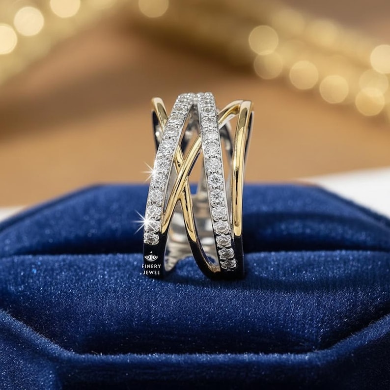 K&ouml;nnte beinhalten: Ein zweifarbiger Ring mit einem Kreuzmuster. Der Ring hat ein Band aus kleinen, klaren Steinen, die in einem silberfarbenen Metall gefasst sind, mit goldfarbenen Akzenten. Der Ring wird auf einer dunkelblauen Samtoberfl&auml;che pr&auml;sentiert.