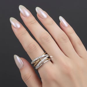 K&ouml;nnte beinhalten: Ein mehrreihiger Ring mit Gold- und Silberdesign. Der Ring hat ein Band aus kleinen, klaren Steinen und verschlungenen Gold- und Silberb&auml;ndern. Der Ring wird an einem Finger vor dunklem Hintergrund getragen.