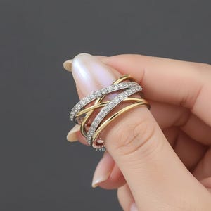 K&ouml;nnte beinhalten: Ein zweifarbiges Ring mit einem Kreuzmuster. Der Ring hat ein goldenes Band, das mit B&auml;ndern verschlungen ist, die mit kleinen, klaren Edelsteinen besetzt sind. Der Ring wird vom Finger einer Person gehalten, was das komplizierte Design und den Glanz des Schmucks zeigt.