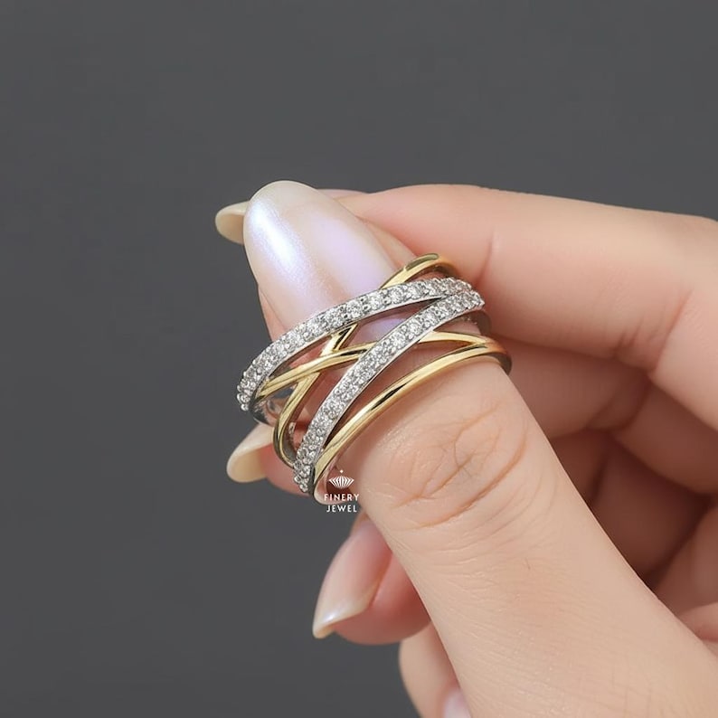 K&ouml;nnte beinhalten: Ein zweifarbiges Ring mit einem Kreuzmuster. Der Ring hat ein goldenes Band, das mit B&auml;ndern verschlungen ist, die mit kleinen, klaren Edelsteinen besetzt sind. Der Ring wird vom Finger einer Person gehalten, was das komplizierte Design und den Glanz des Schmucks zeigt.