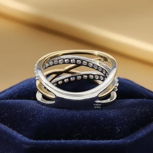 K&ouml;nnte beinhalten: Ein zweifarbiges Ring mit einem Kreuzmuster. Der Ring hat ein goldenes Band, das mit einem silbernen Band verschlungen ist, beide mit kleinen, funkelnden Steinen verziert. Der Ring liegt auf einer dunkelblauen Samtoberfl&auml;che.