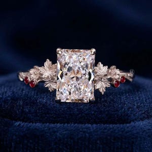 May include: A silver-toned ring featuring a large, rectangular, faceted clear center stone. The band is detailed with intricate metal leaf motifs on both sides, accented by small, round, deep red stones embedded within the foliage design.