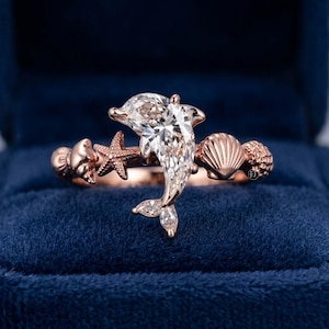 May include: A rose gold ring featuring a central marquise-cut clear gemstone shaped like a dolphin. The band is decorated with a small starfish, a scallop shell, and a textured sea shell motif, all crafted in a polished rose gold metal finish.
