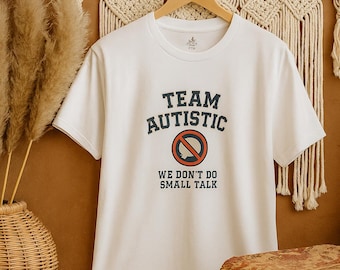 Könnte beinhalten: Weißes T-Shirt mit dem Text "TEAM AUTISTIC" in Marineblau, mit einem roten Kreis und einem durchgestrichenen Symbol. Darunter steht "WE DON'T DO SMALL TALK". Das Shirt hängt an einem Holzkleiderbügel.