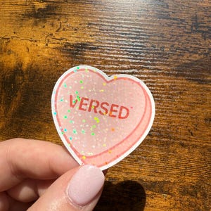May include: A heart-shaped sticker with the word "VERSED" in red. The sticker has a pink glittery interior and a red outline. The sticker is held by a person.