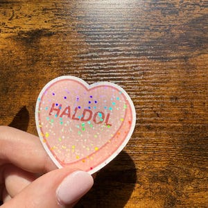 May include: A heart-shaped sticker with the word "HALDOL" in red. The sticker has a pink background with iridescent glitter and a white border. The sticker is held by a person.
