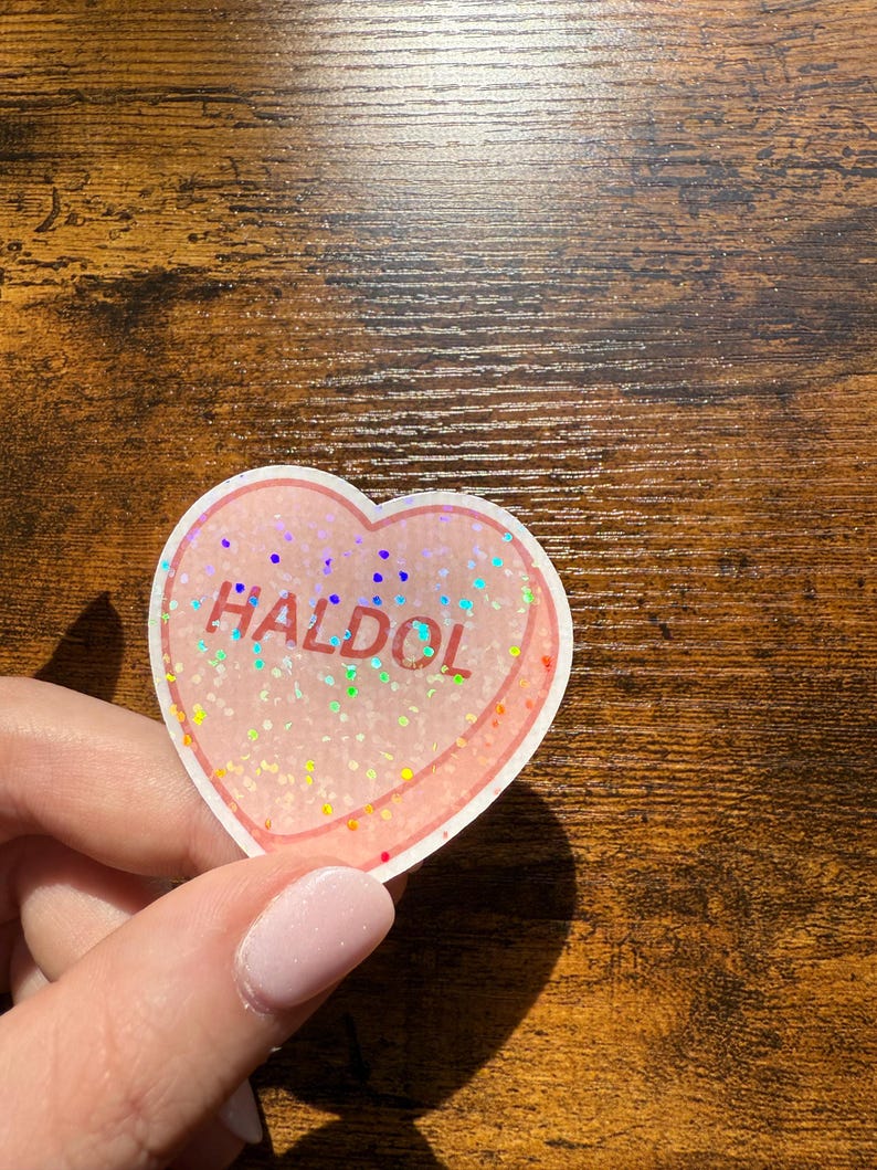 May include: A heart-shaped sticker with the word "HALDOL" in red. The sticker has a pink background with iridescent glitter and a white border. The sticker is held by a person.