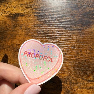 May include: A heart-shaped sticker with the word "PROPOFOL" in purple letters. The sticker has a pink border and is filled with iridescent glitter. The sticker is held by a person.