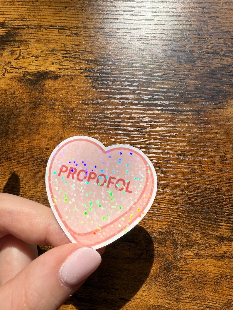 May include: A heart-shaped sticker with the word "PROPOFOL" in purple letters. The sticker has a pink border and is filled with iridescent glitter. The sticker is held by a person.