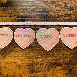 May include: Four heart-shaped stickers with pink glitter and white borders. Each sticker has a different word: PROPOFOL, KETAMINE, HALDOL, and VERSED. A measuring tape is above the stickers.