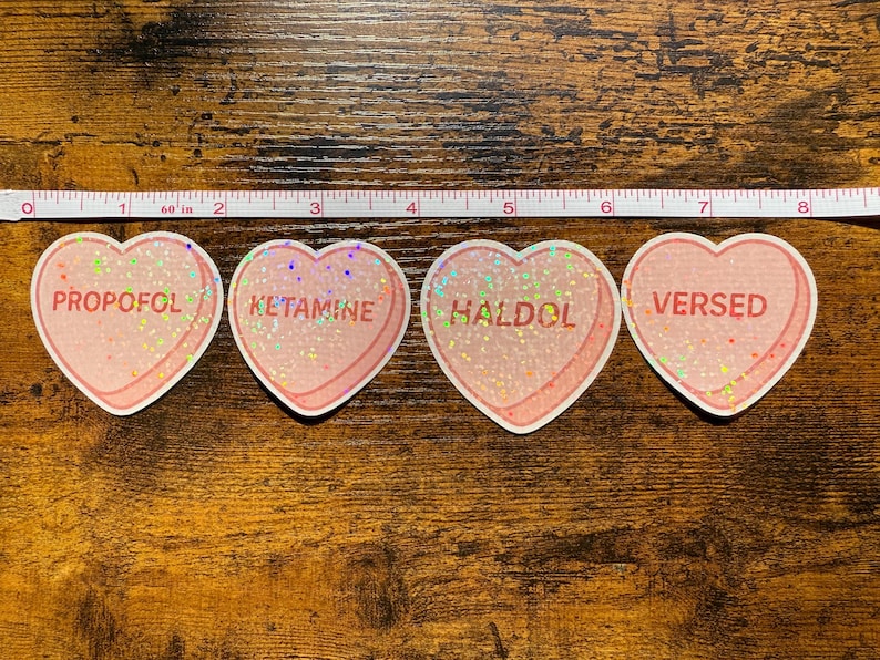 May include: Four heart-shaped stickers with pink glitter and white borders. Each sticker has a different word: PROPOFOL, KETAMINE, HALDOL, and VERSED. A measuring tape is above the stickers.