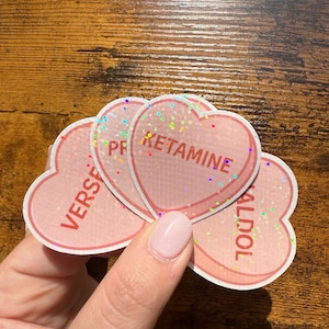 May include: Pink heart-shaped stickers with glitter accents. Each sticker has a different word printed on it: "KETAMINE", "VERS", and "HALDOL". The stickers are held by a person.