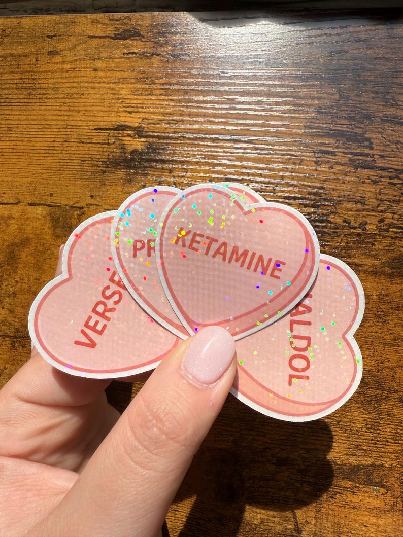 May include: Pink heart-shaped stickers with glitter accents. Each sticker has a different word printed on it: "KETAMINE", "VERS", and "HALDOL". The stickers are held by a person.