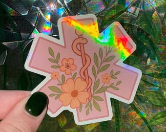 May include: A pink sticker in the shape of a medical star with a floral design. The sticker features a caduceus symbol, peach-colored flowers, and green leaves. The sticker has a holographic, rainbow-colored border.