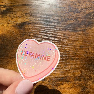 May include: A heart-shaped sticker with the word "KETAMINE" in red and glittery text. The sticker has a pink border and a clear, glittery centre. The sticker is held by a person.