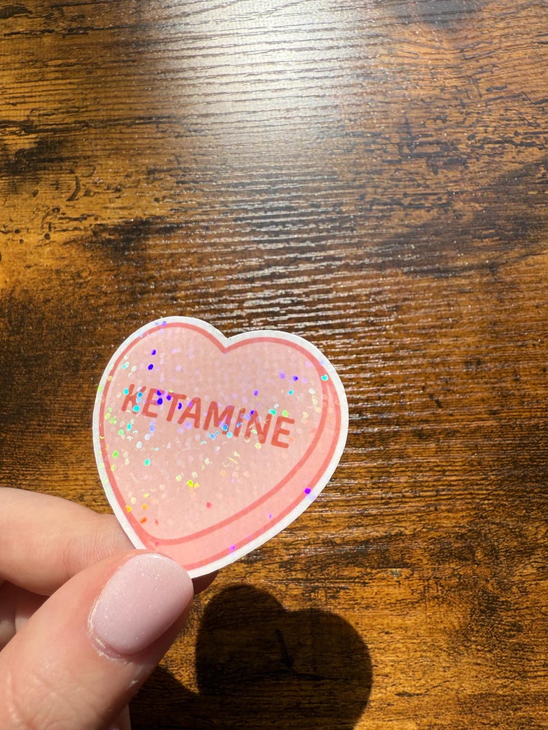 May include: A heart-shaped sticker with the word "KETAMINE" in red and glittery text. The sticker has a pink border and a clear, glittery centre. The sticker is held by a person.