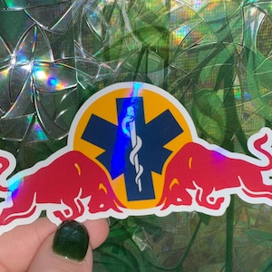 Puede incluir: Una pegatina con dos toros rojos flanqueando un círculo amarillo con una cruz azul y un símbolo médico. La pegatina tiene un efecto holográfico, con colores iridiscentes visibles.