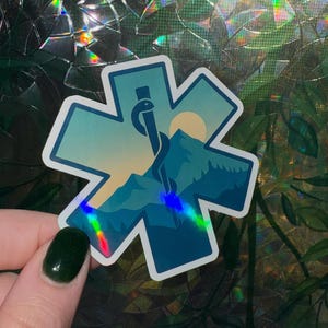May include: A sticker in the shape of a blue star of life with a mountain and sun scene. A medical symbol with a snake and staff is in the center. The sticker has a holographic effect.