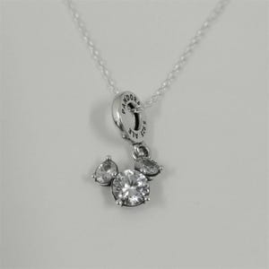 May include: Silver-toned pendant necklace featuring a charm with a Mickey Mouse-inspired design. The charm has a large, round, clear gemstone at the center, with smaller, pear-shaped gemstones on either side. The necklace has a delicate chain.