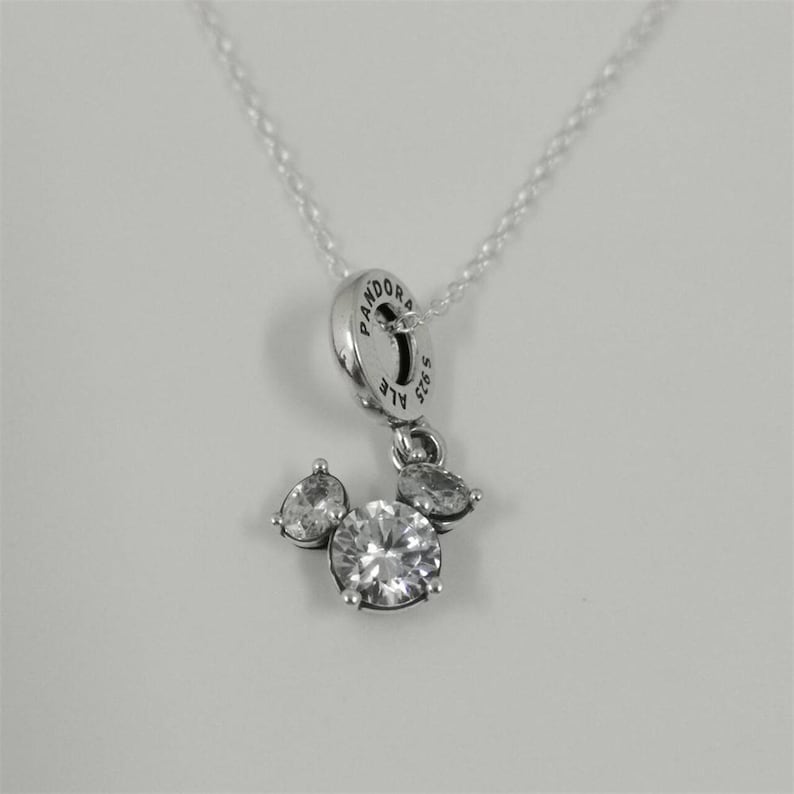 May include: Silver-toned pendant necklace featuring a charm with a Mickey Mouse-inspired design. The charm has a large, round, clear gemstone at the center, with smaller, pear-shaped gemstones on either side. The necklace has a delicate chain.