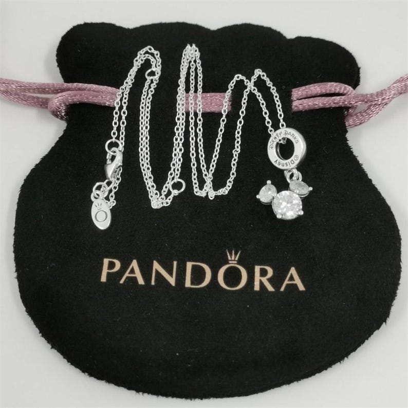 May include: A silver necklace with a Mickey Mouse pendant, displayed on a black Pandora bag. The pendant features a large, round, clear stone and two smaller stones, with the Disney logo. The bag has a pink drawstring.
