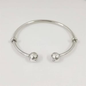 May include: A silver open bangle bracelet. The bracelet has a rounded, open design with a ball-shaped end on each side. The balls have the word "Pandora" engraved on them. The bracelet is a jewelry item.