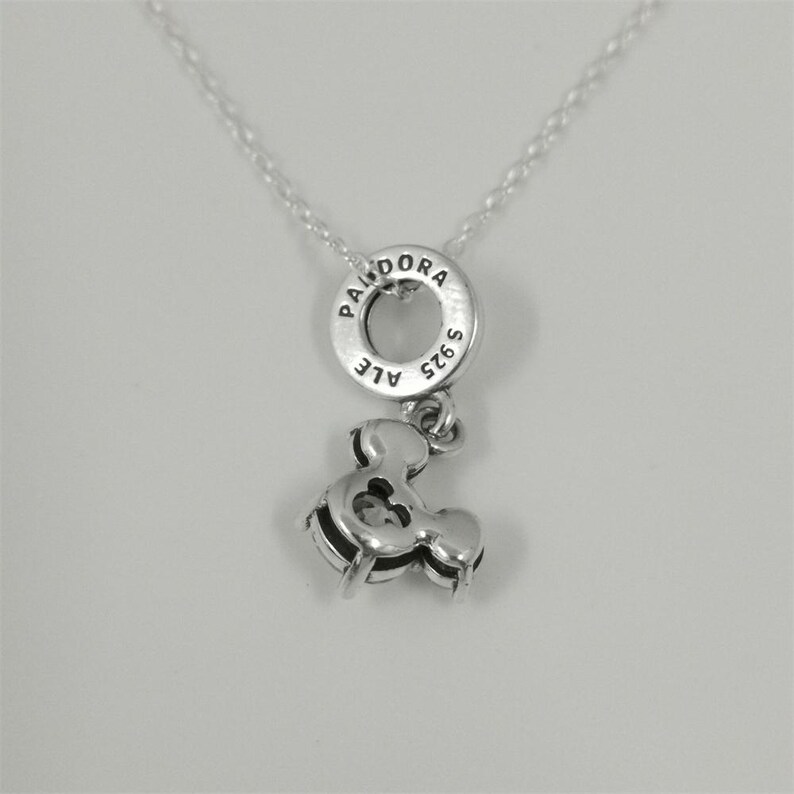May include: A silver necklace featuring a Pandora charm. The charm includes a circular pendant with the brand name and a smaller ampersand charm. The necklace is displayed against a white background.