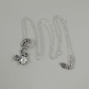 May include: A silver necklace featuring a Mickey Mouse pendant with clear crystal accents. The pendant is attached to a delicate chain. The necklace has a tag that reads "S925 ALE". The necklace is displayed on a white surface.