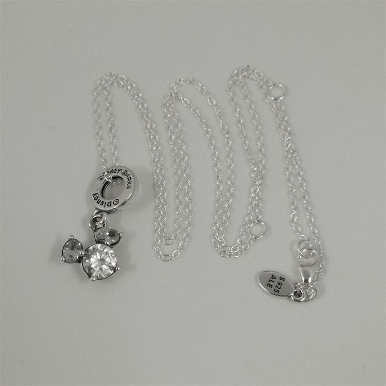May include: A silver necklace featuring a Mickey Mouse pendant with clear crystal accents. The pendant is attached to a delicate chain. The necklace has a tag that reads "S925 ALE". The necklace is displayed on a white surface.