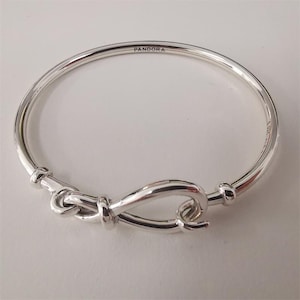 May include: A silver Pandora bangle bracelet featuring a knot-shaped clasp. The bracelet has the brand name "Pandora" engraved on the top. The clasp is a distinctive knot design, adding a stylish touch.